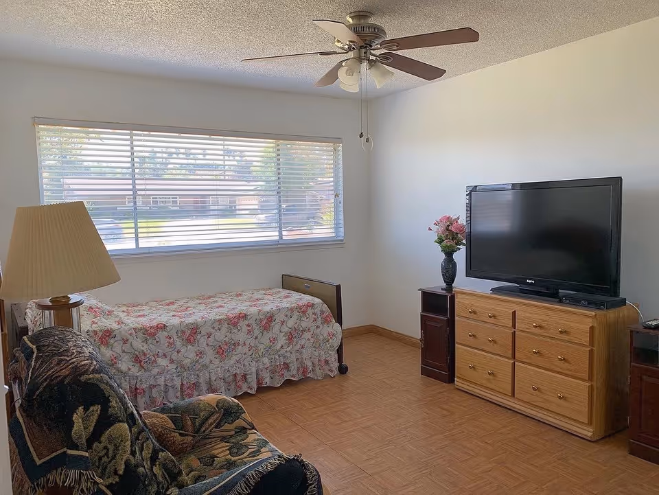 A simple bedroom with a single bed covered in a floral bedspread, a wooden dresser with a flat-screen TV on top, a small side table with a vase of flowers, a cushioned armchair with a patterned throw, a large window with horizontal blinds, and a ceiling fan with lights.