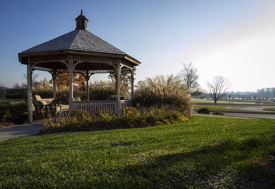 A wooden gazebo with a shingled roof situated in a landscaped garden area with green grass, ornamental grasses, and some trees in the background under a clear sky.