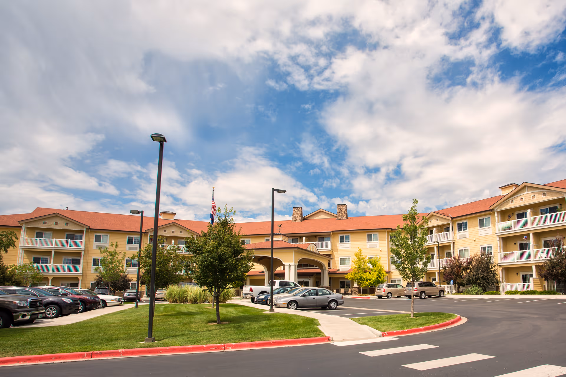 Exterior view of a senior living facility building with a red roof, beige siding, balconies, and a covered entrance. There are several cars parked in the parking lot, green grass, trees, and a partly cloudy blue sky overhead.