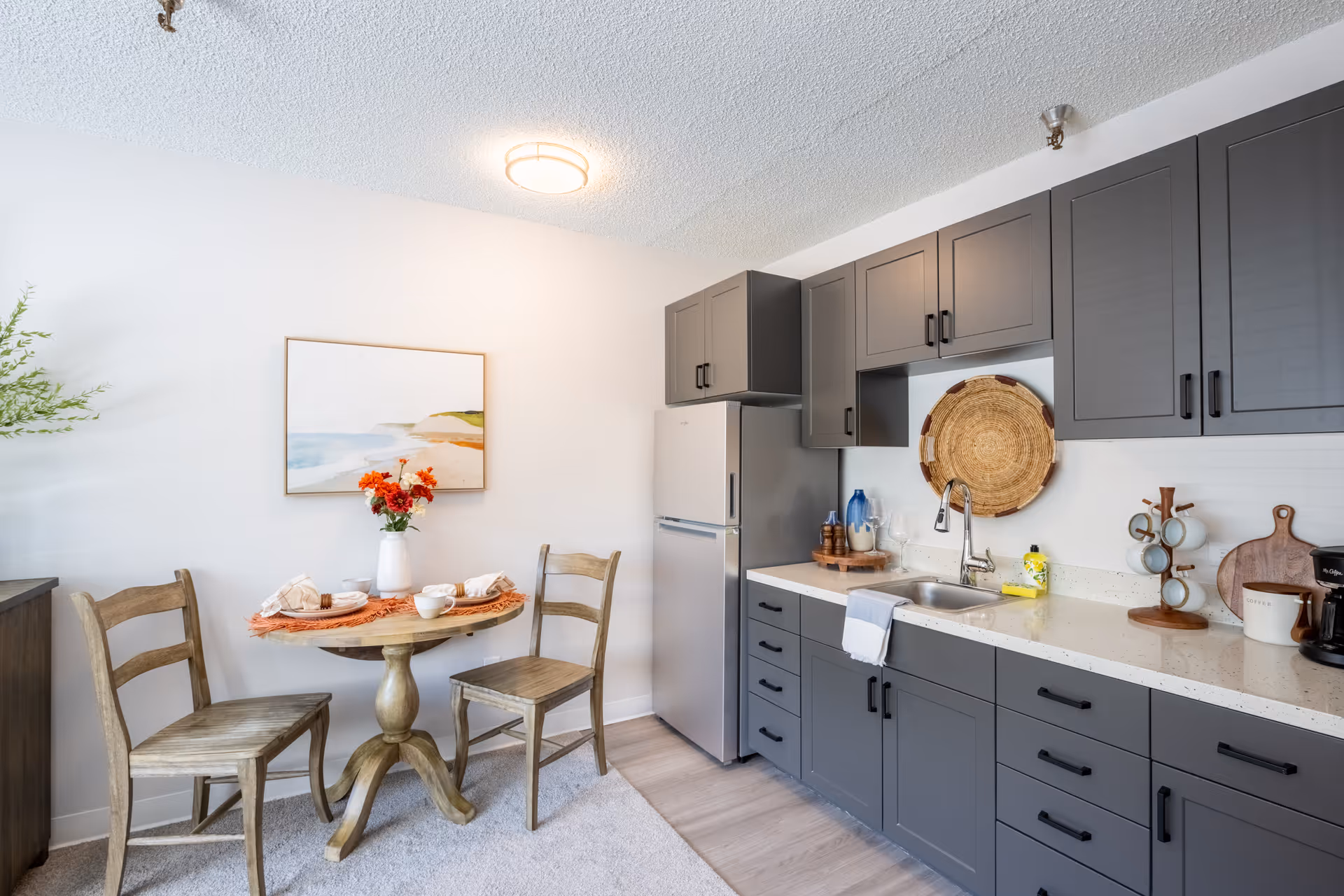 Compact kitchen with gray cabinets, a refrigerator and sink, and a small round dining table set with two wooden chairs.