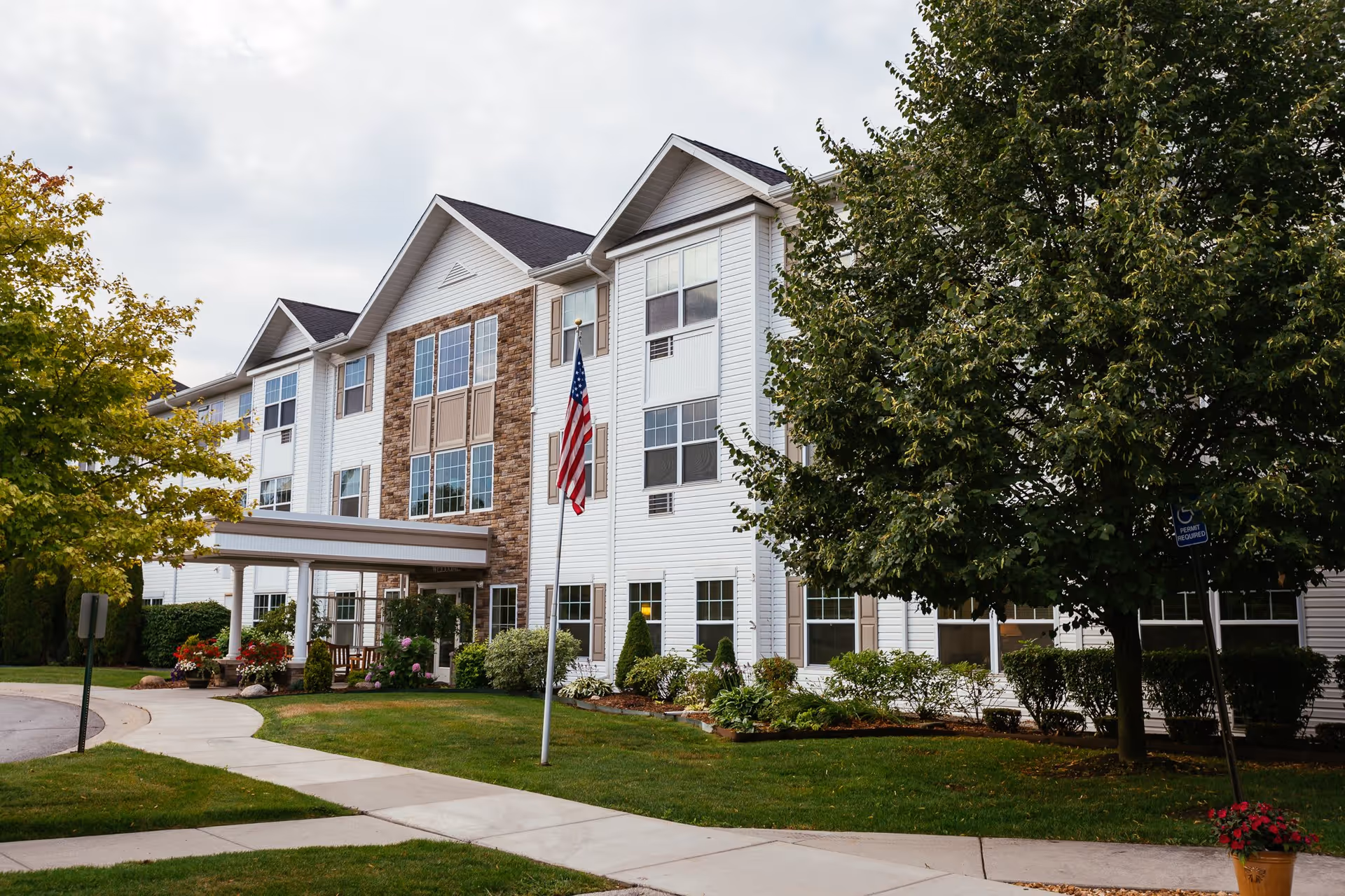 Exterior view of a multi-story senior living facility with white siding and stone accents, surrounded by green trees and landscaping. A sidewalk curves in front of the building, and an American flag is displayed on a flagpole near the entrance.