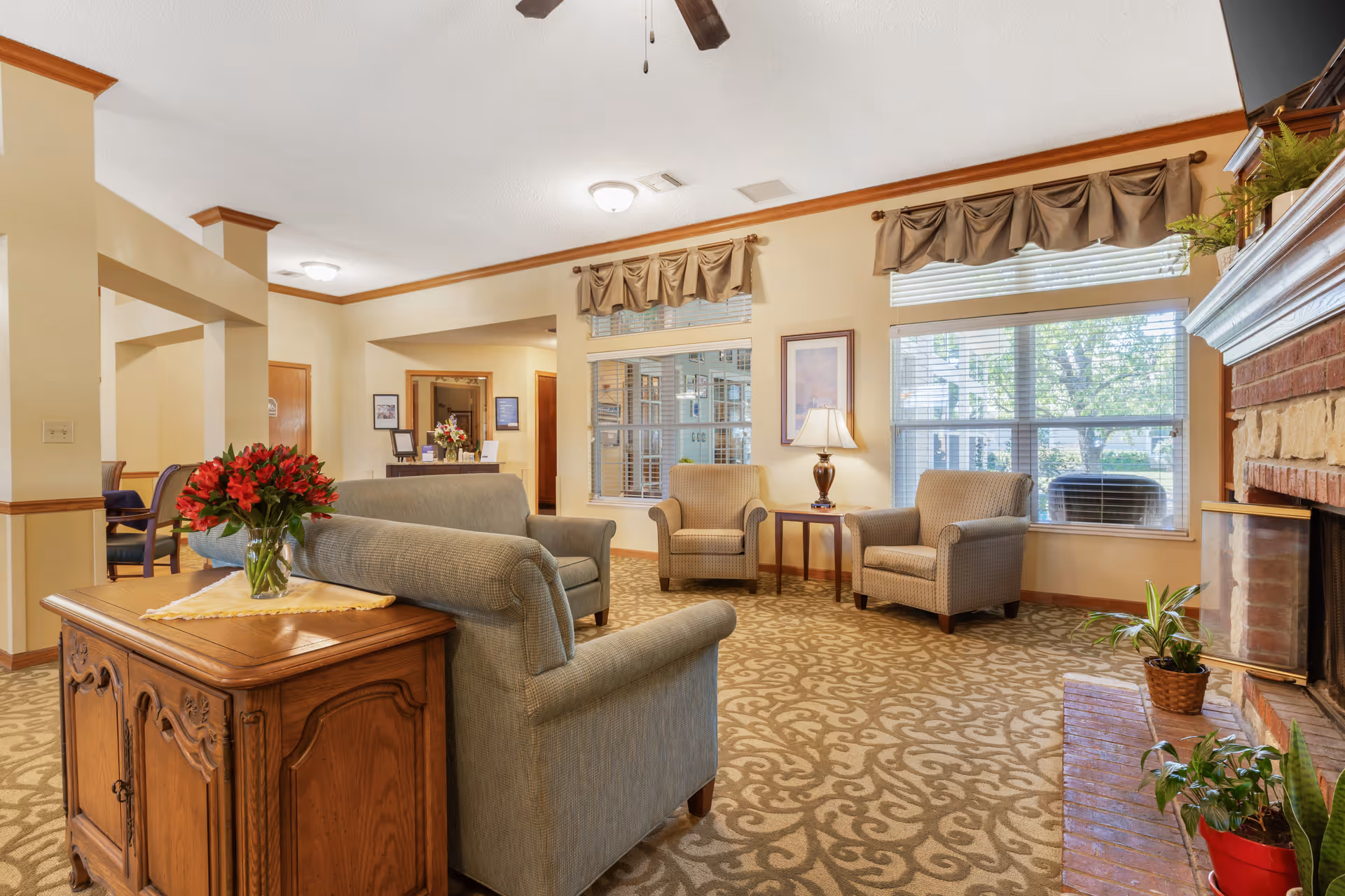 A cozy living room area in a senior living facility with beige patterned carpet, two armchairs, a loveseat, a wooden side table with a vase of red flowers, a lamp on a small table between the armchairs, large windows with blinds and valances, and a brick fireplace with potted plants on the hearth.