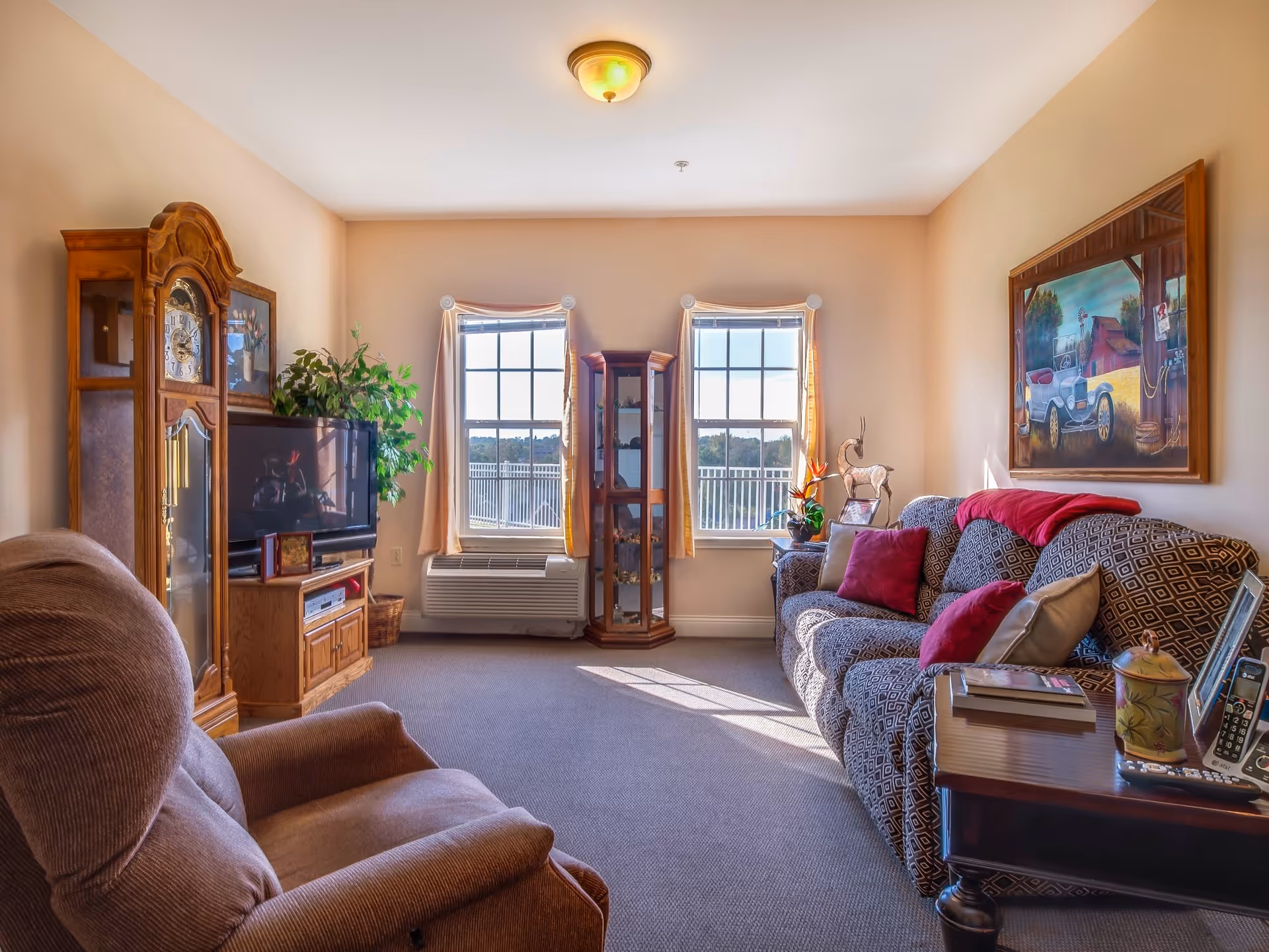 A cozy living room in a senior living facility with a patterned sofa adorned with red and beige pillows, a brown recliner chair, a wooden grandfather clock, a TV on a wooden stand, a glass display cabinet between two windows with beige curtains, and a side table with books, a phone, and decorative items. A colorful painting hangs on the wall above the sofa.