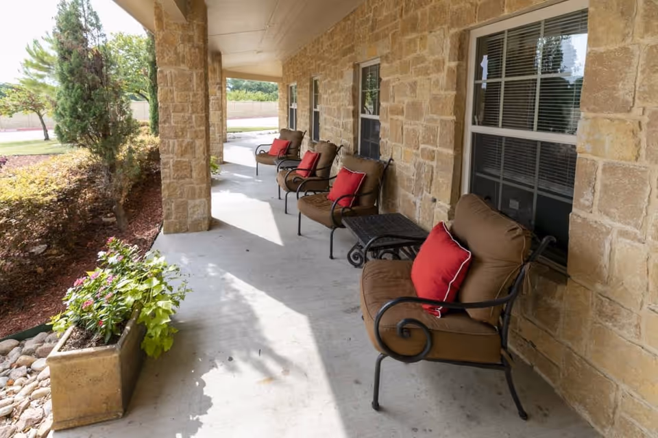 Covered outdoor patio area with four cushioned chairs, each with a red pillow, arranged along a stone wall with windows. There is a small table between two of the chairs and a planter with green plants and flowers at the edge of the patio. Trees and landscaping are visible beyond the patio.