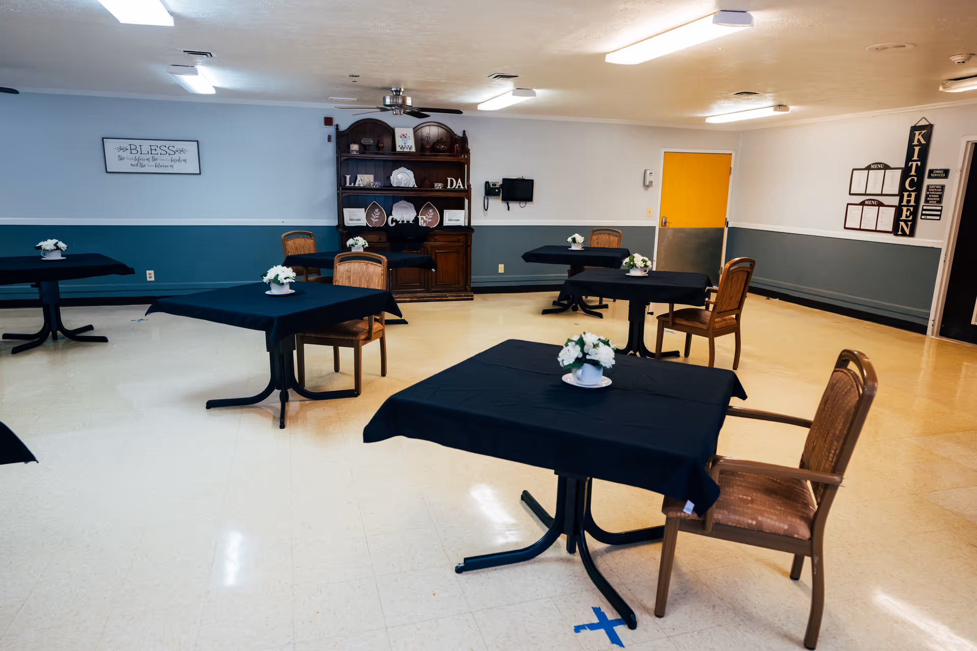 A dining room with several square tables covered with black tablecloths, each adorned with a small white floral centerpiece. Wooden chairs are placed around the tables. The walls are painted in two tones, with a blue lower half and white upper half. A wooden hutch with decorative items is against the far wall, and a yellow door is visible on the right side. A sign on the wall reads 'KITCHEN' with menus displayed underneath.