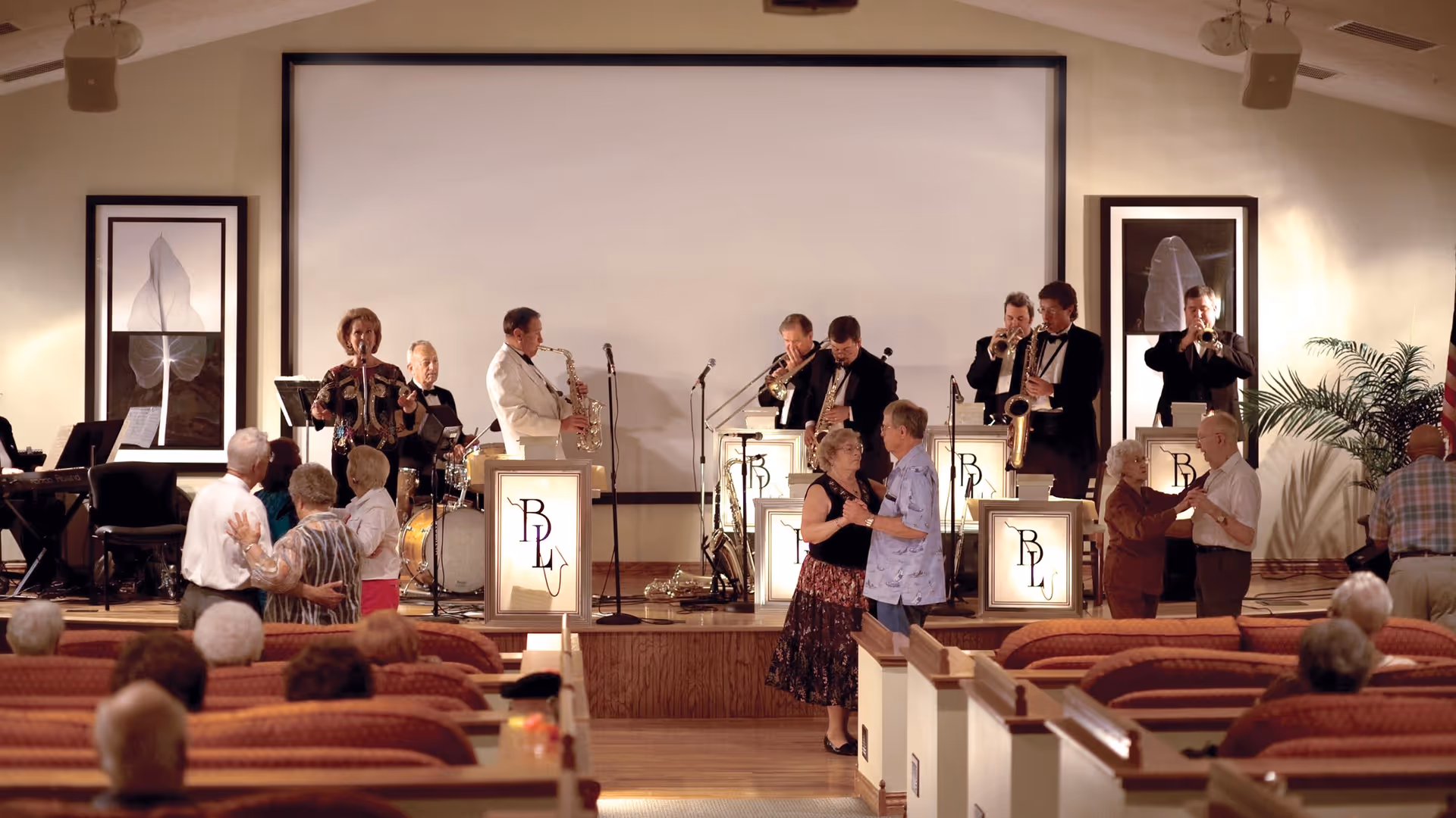 A live band performs on a stage in front of a large white screen, with elderly couples dancing in front of the stage and seated audience members watching. The band includes musicians playing saxophone, trumpet, trombone, drums, and keyboard, with a female vocalist. The setting appears to be a community or event room with framed artwork on the walls and potted plants.