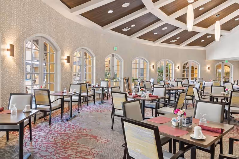 A spacious dining room with multiple tables and chairs arranged neatly. Each table is set with placemats, napkins, cups, and small flower arrangements. The room features large arched windows allowing natural light to fill the space, patterned carpet, and a coffered ceiling with recessed lighting.