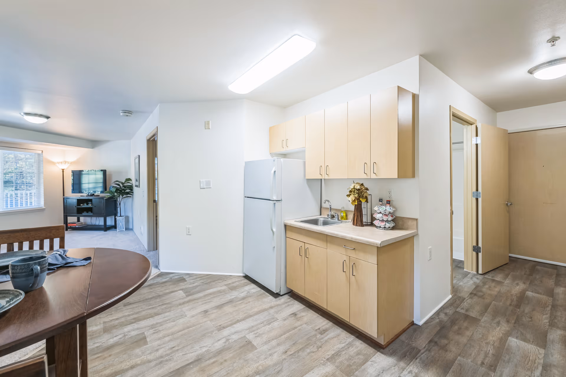 Interior view of a senior living facility apartment showing a small kitchen area with light wood cabinets, a white refrigerator, and a countertop with a sink and decorative items. Adjacent to the kitchen is a dining table with place settings. In the background, there is a living area with a TV on a stand, a floor lamp, and a window with blinds. The flooring is wood-style laminate, and the walls are painted white.