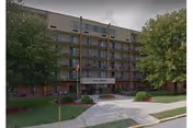 Exterior view of a multi-story senior living facility named Southpoint at Jameson, featuring balconies on each floor, a main entrance with a covered drop-off area, surrounded by trees and landscaped greenery.