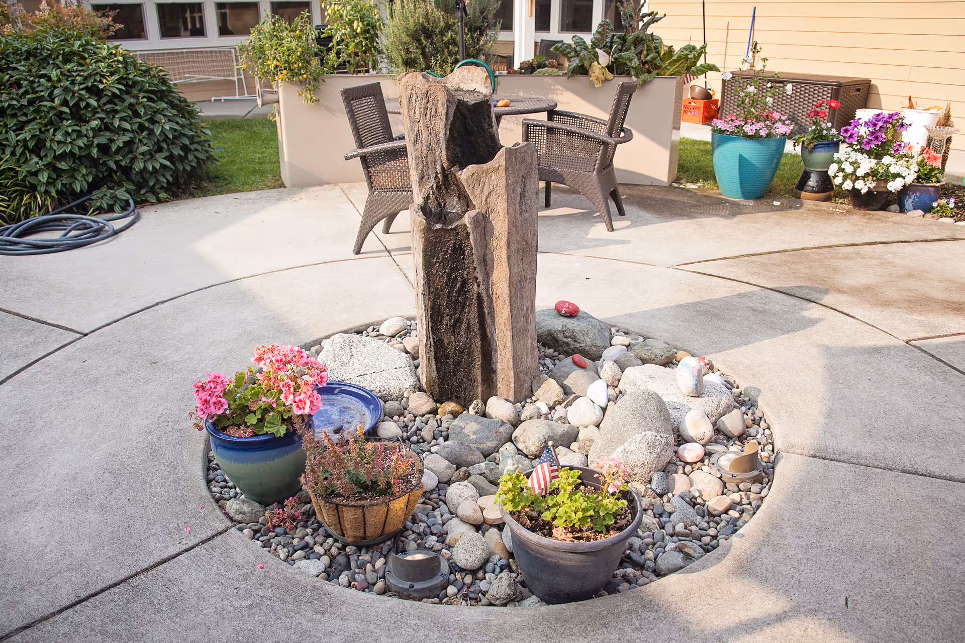 Circular outdoor courtyard with a central rock planter and wooden fountain surrounded by potted flowers and patio chairs.