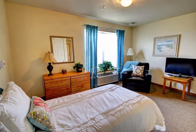A cozy bedroom in a senior living facility featuring a neatly made bed with white bedding and decorative pillows, a wooden dresser with a lamp and plants, a window with blue curtains, a black armchair with a cushion and throw blanket, a floor lamp, a framed beach-themed picture on the wall, and a flat-screen TV on a wooden stand.