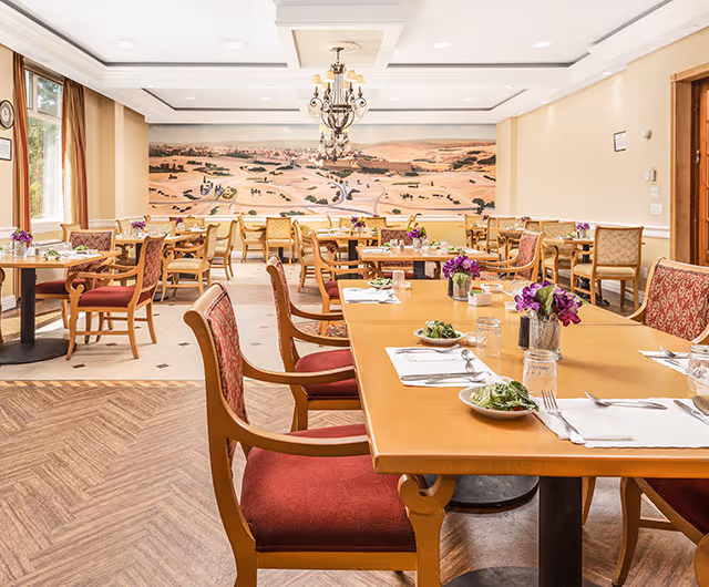 A bright dining room with multiple wooden tables and chairs, each table set with plates of salad, glasses, and silverware. The chairs have red and beige upholstery. The room features a large mural on the far wall depicting a scenic landscape. There are windows on the left side letting in natural light and a chandelier hanging from the ceiling.
