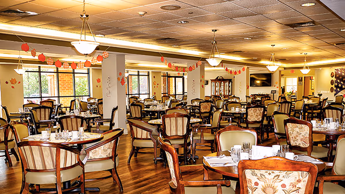 A spacious dining room with multiple round wooden tables and upholstered chairs arranged neatly. The room is decorated with autumn-themed garlands and wall decals featuring leaves and pumpkins. Large windows allow natural light to brighten the space, and ceiling lights provide additional illumination. The floor is wooden, and the room has a warm, inviting atmosphere.