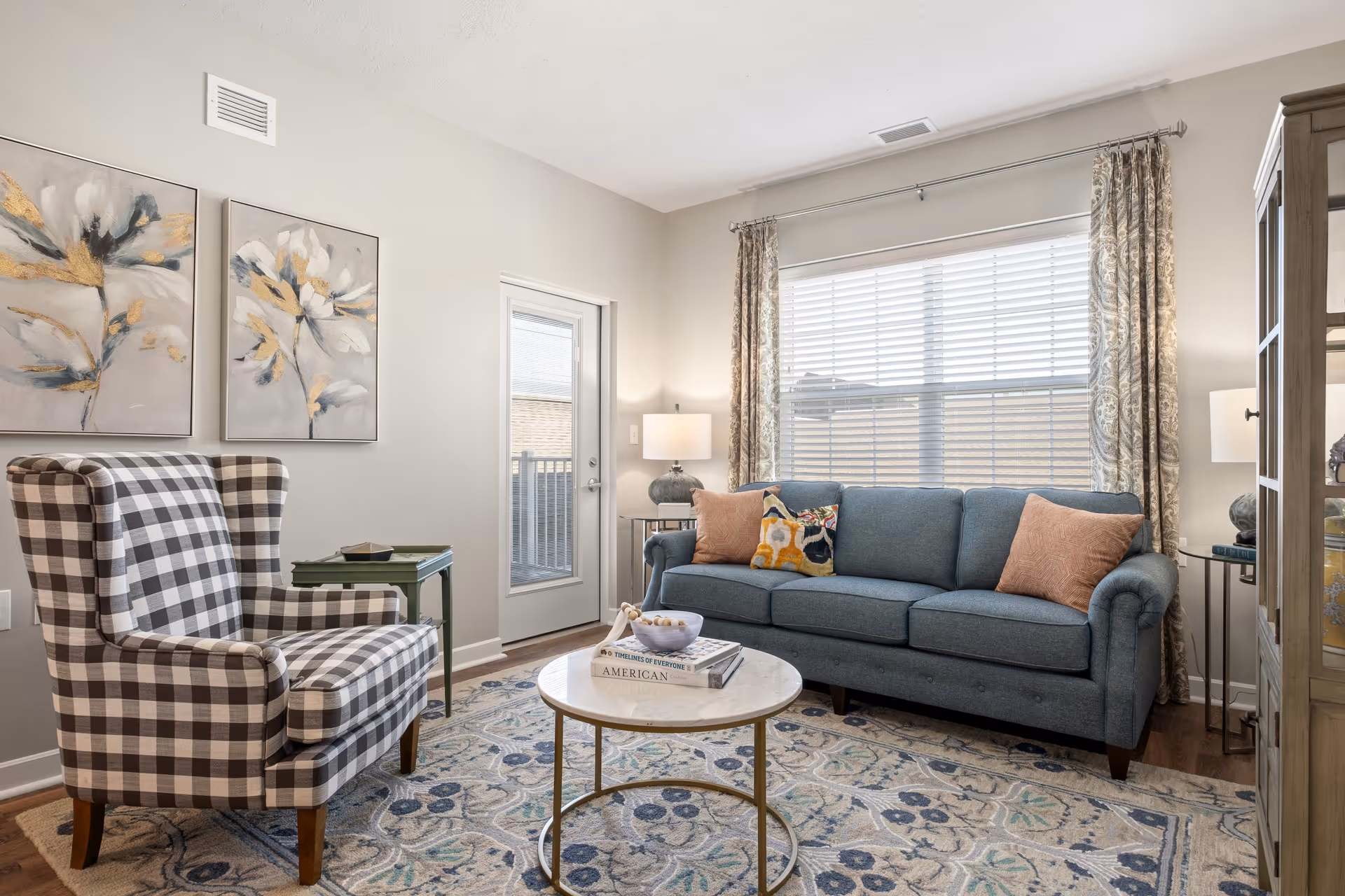 Cozy living room with a blue upholstered sofa, plaid armchair, round marble-top coffee table, and a large window with drapes.