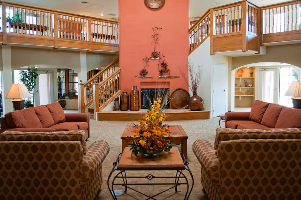A spacious senior living facility common area with comfortable seating including patterned armchairs and solid-colored sofas arranged around a wooden coffee table with a floral centerpiece. The room features a large red fireplace with decorative items on the mantel and a wooden staircase with railings leading to an upper level balcony. There are lamps on side tables and large windows letting in natural light.