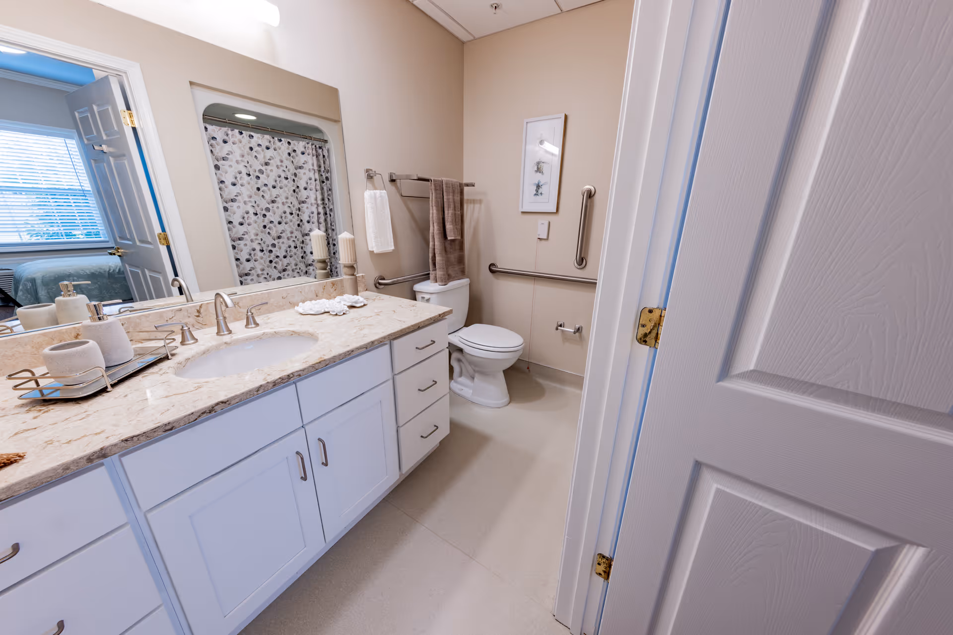 Well-lit accessible bathroom with a white vanity and marble countertop, sink, toilet with grab bars, and a shower with a patterned curtain.