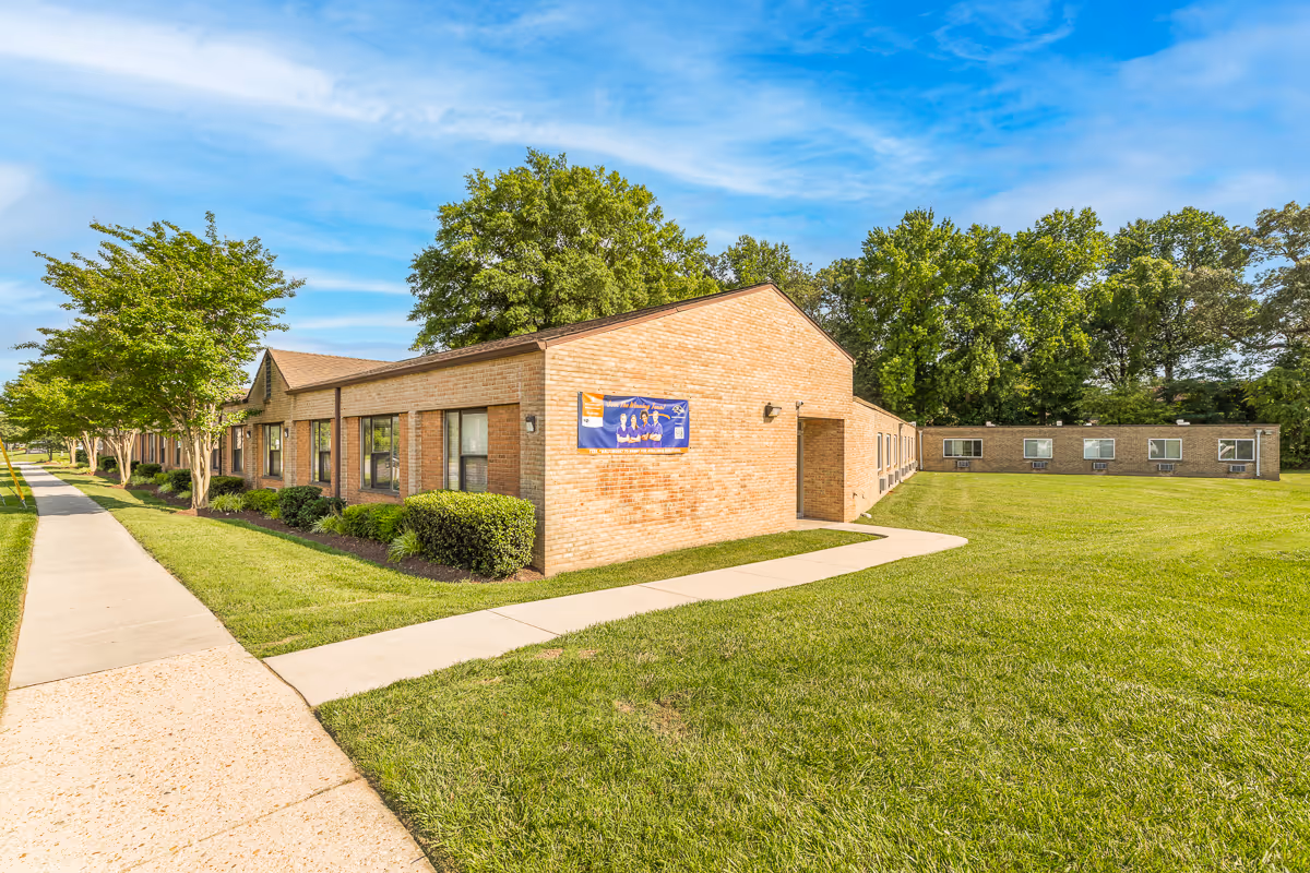 Exterior view of a single-story brick building with multiple windows, surrounded by green grass and trees under a blue sky with some clouds. A sidewalk runs alongside the building, and there is a banner on the side wall.