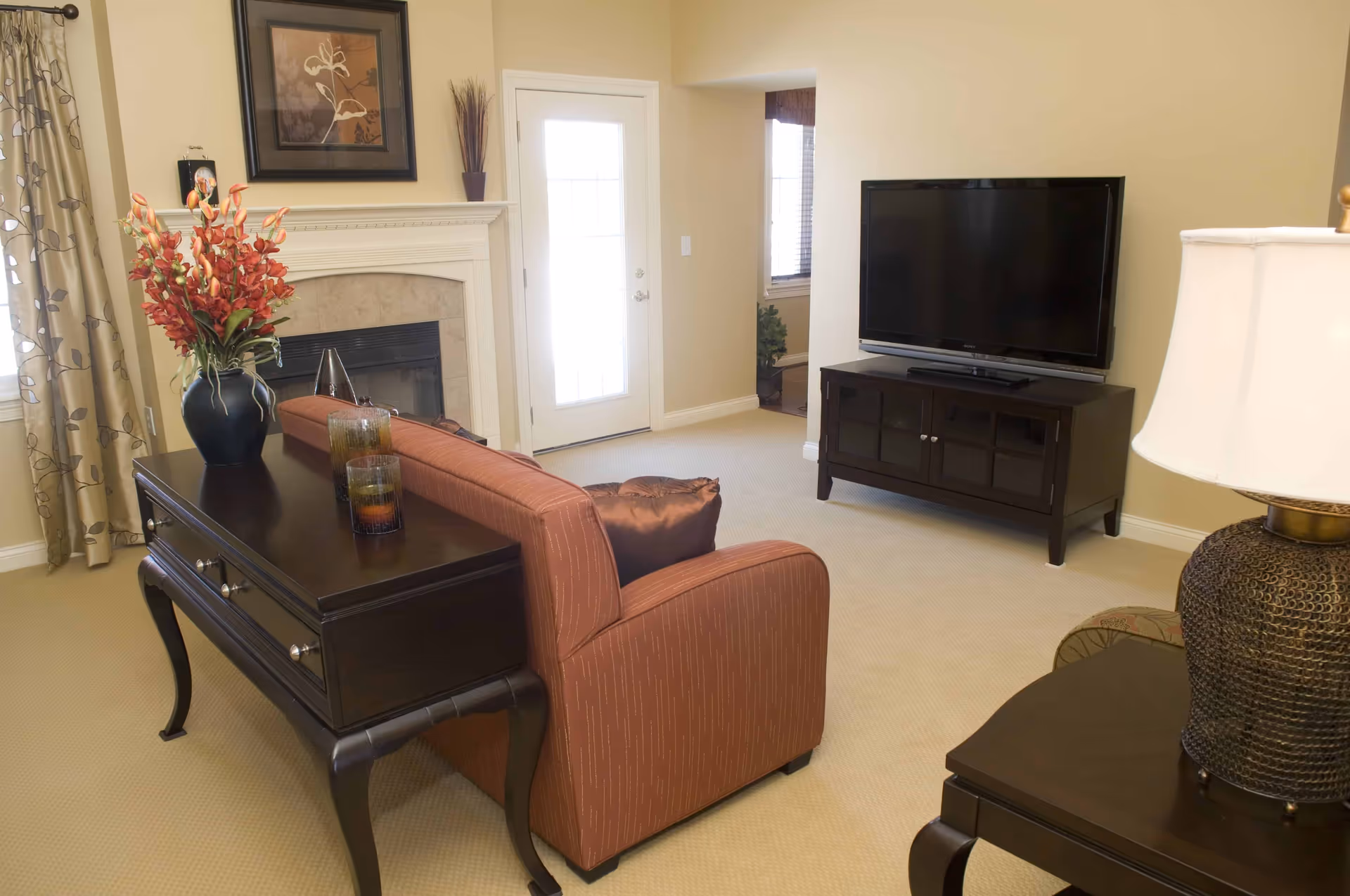 A well-lit living room with a sofa, flat-screen TV on a stand, fireplace, and decorative tables with a lamp and vase.