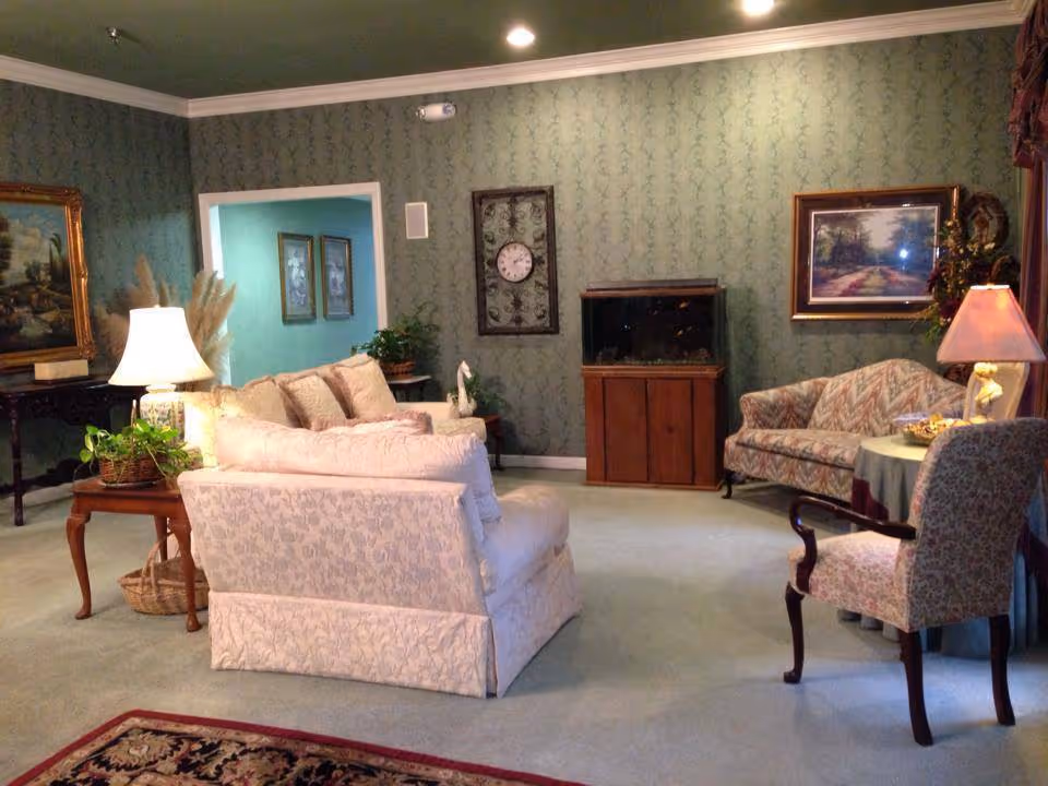 A cozy living room with floral patterned sofas and armchairs arranged around a carpet. The walls are covered with green wallpaper and decorated with framed paintings and a clock. There is a wooden cabinet with a fish tank on top, a side table with a lamp, and various plants adding a touch of greenery.