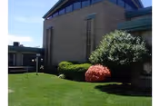 Exterior view of a brick building with a well-maintained lawn and bushes, including a large green bush and a smaller red bush, under a clear blue sky.