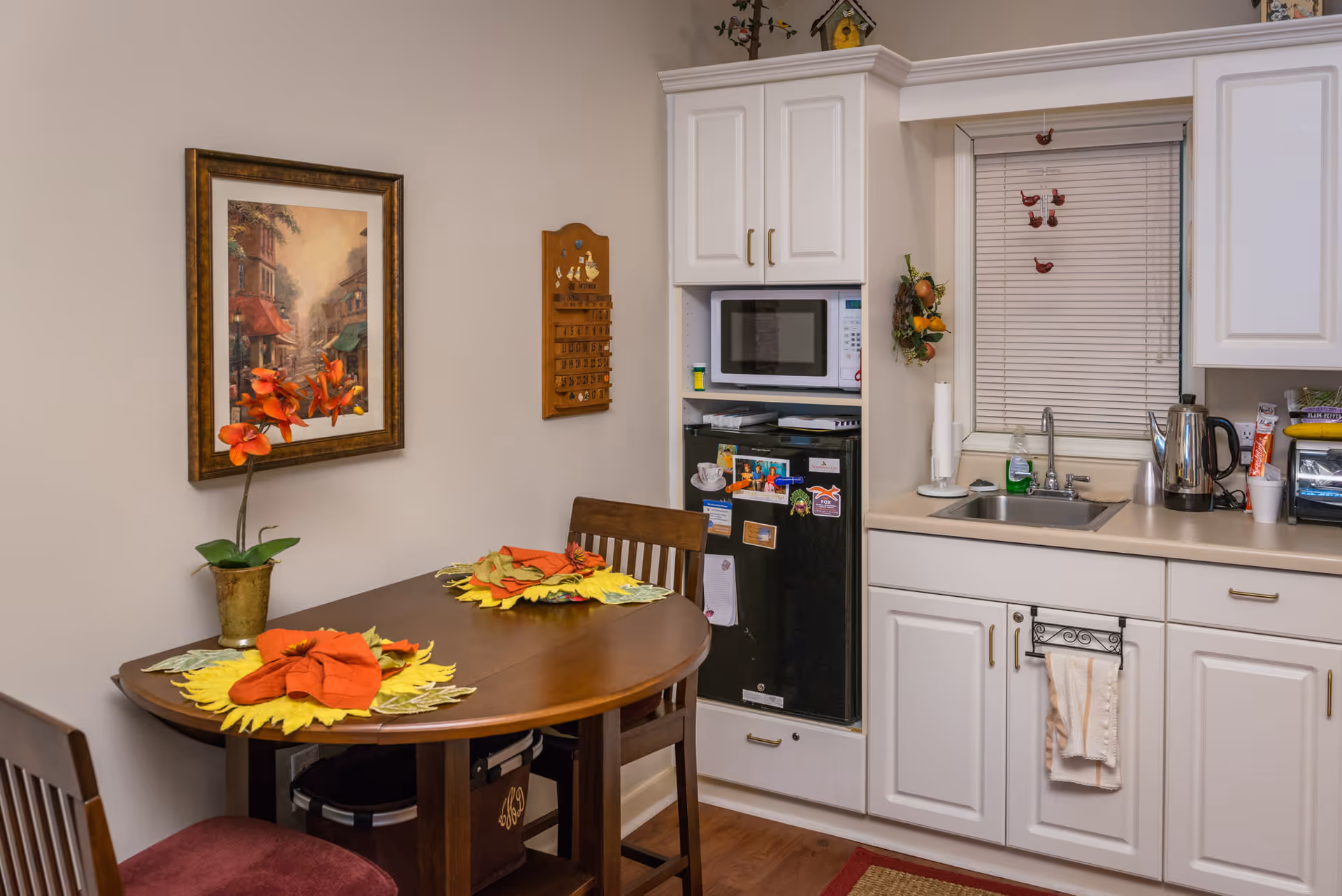 Small kitchen and dining nook with a wooden table set with floral placemats, white cabinets, sink, microwave, and a mini-fridge.