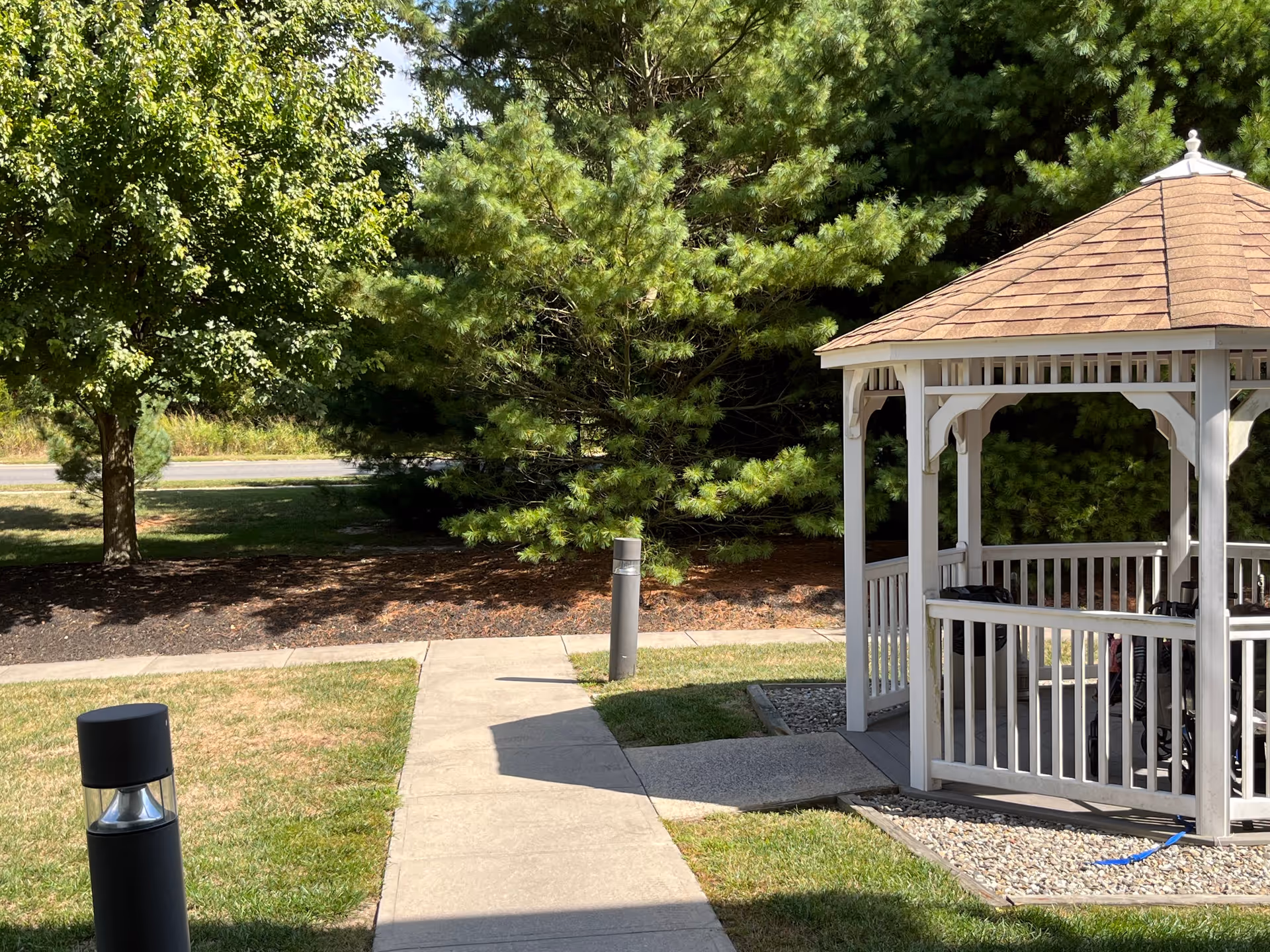 A white wooden gazebo with a shingled roof situated on a grassy area next to a concrete walkway. The background features green trees and a road beyond the landscaped area.