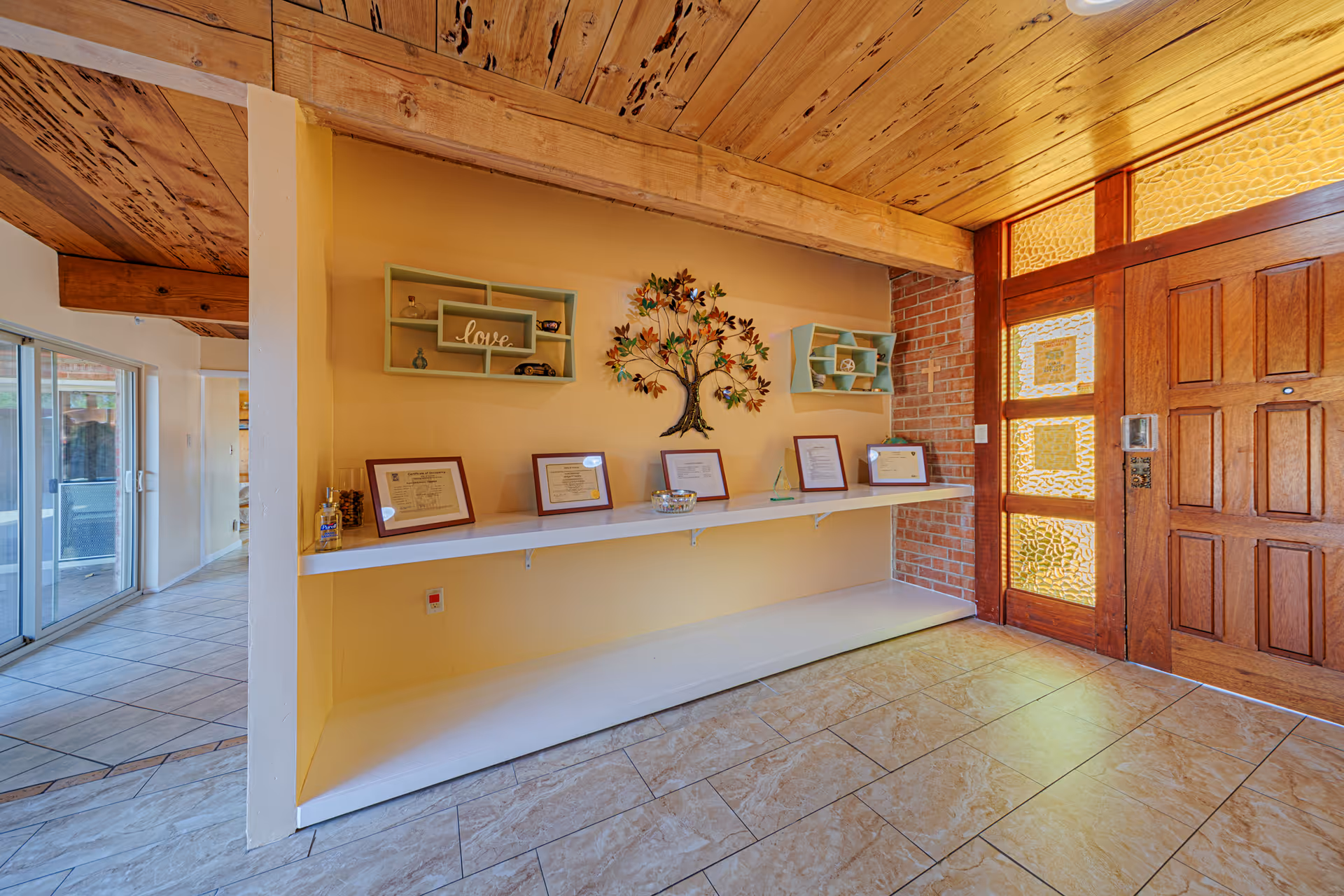 Interior view of an entryway or hallway with a wooden front door featuring textured glass panels. The walls are painted yellow and decorated with a metal tree wall art and two small shelves holding decorative items. A long white floating shelf below displays framed certificates and a glass bowl. The floor is tiled, and the ceiling has exposed wooden beams.