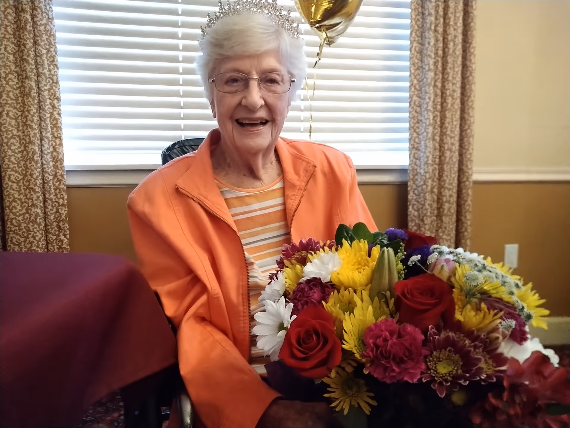 An elderly woman wearing glasses and a tiara, dressed in an orange jacket and striped shirt, smiling while holding a colorful bouquet of flowers. She is seated indoors near a window with blinds and patterned curtains.