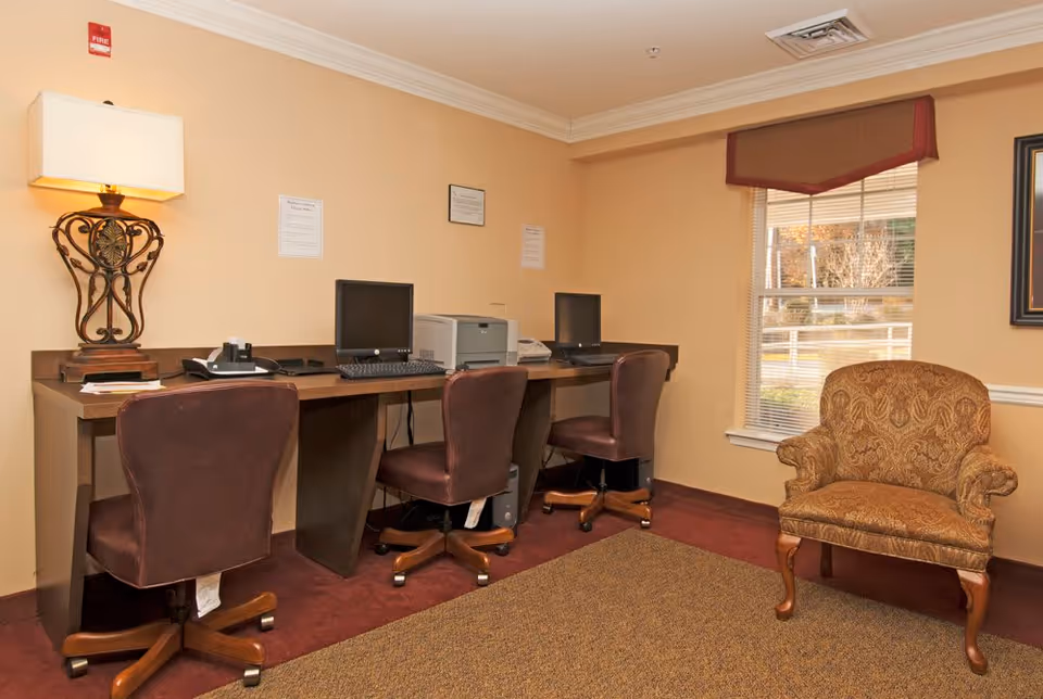 A small office area with three brown leather swivel chairs in front of a long wooden desk. The desk holds two computer monitors, a printer, a telephone, and a decorative lamp. There is a patterned armchair to the right near a window with blinds and a valance. The walls are beige and the carpet is a mix of red and brown tones.