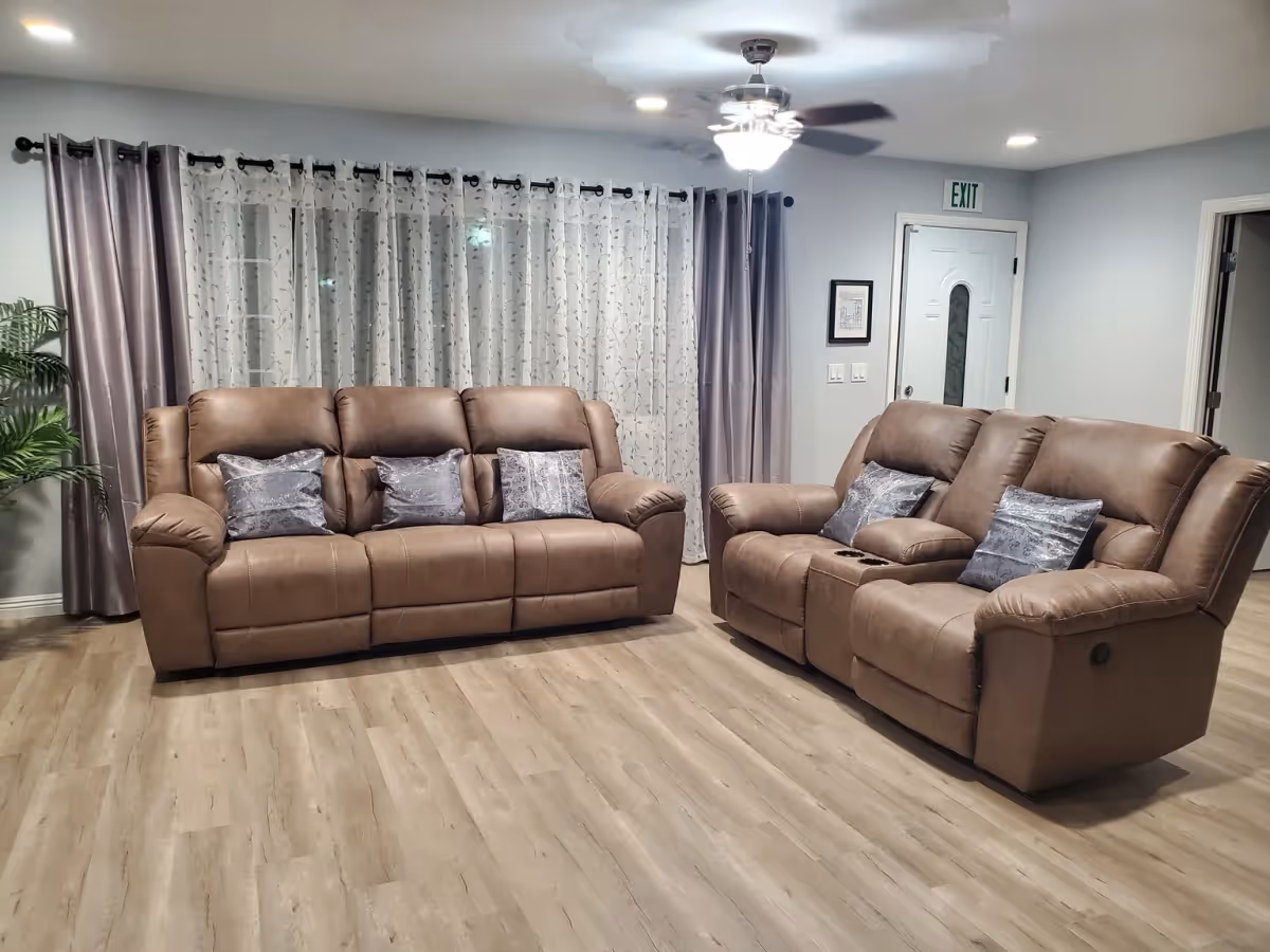 A living room with two brown leather sofas, each with decorative pillows. The room has light wood flooring, sheer white curtains with a floral pattern, and solid gray curtains on the sides. A ceiling fan with lights is on the ceiling, and there is an exit door in the background.