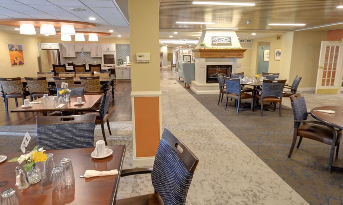 Spacious senior living dining room with multiple set tables and chairs, a central fireplace, and an open kitchen area in the background.