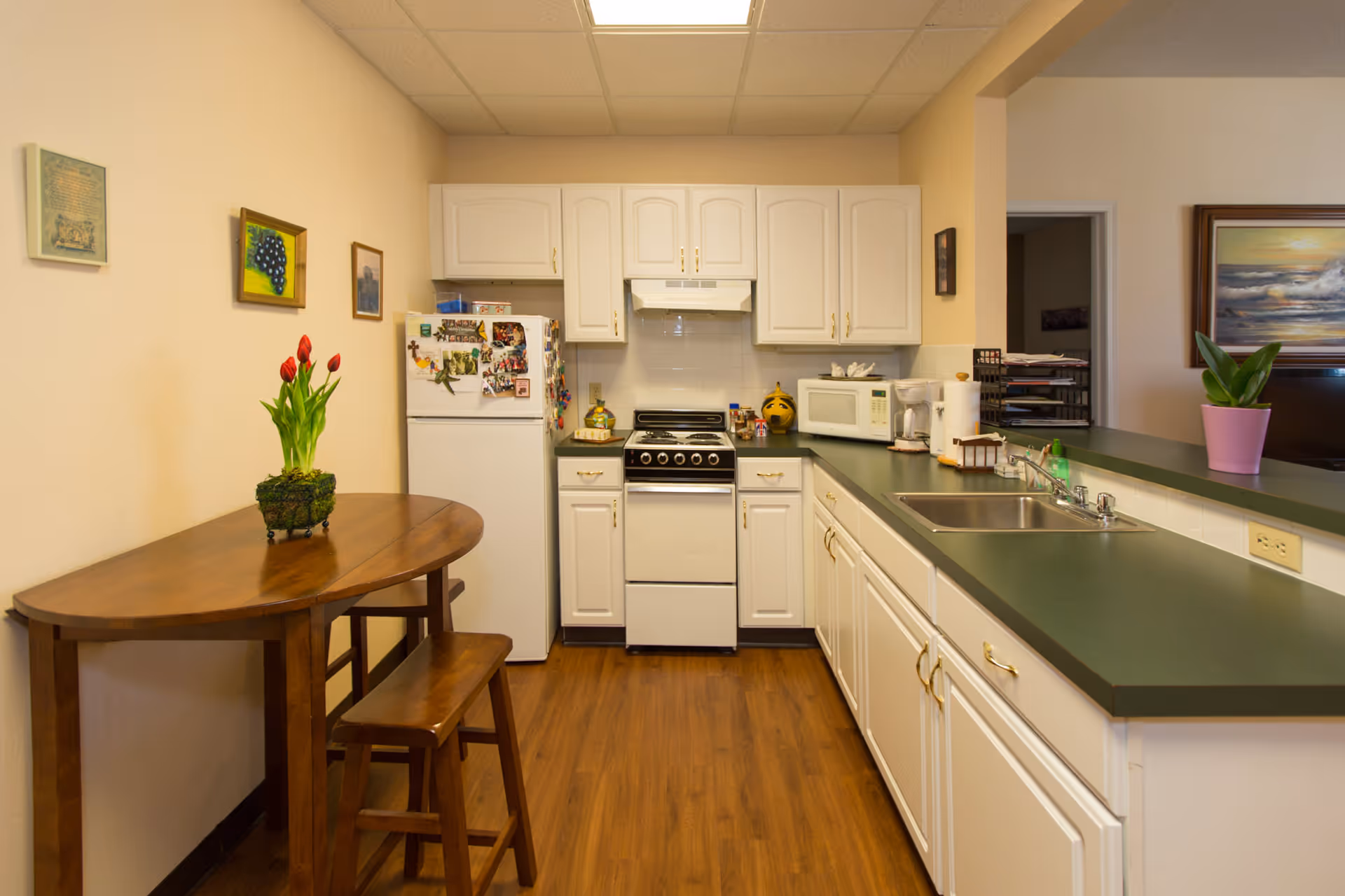 Cozy kitchen with white cabinets, green countertops, a stove and refrigerator, a sink on the right, and a small wooden table with a potted tulip on the left.