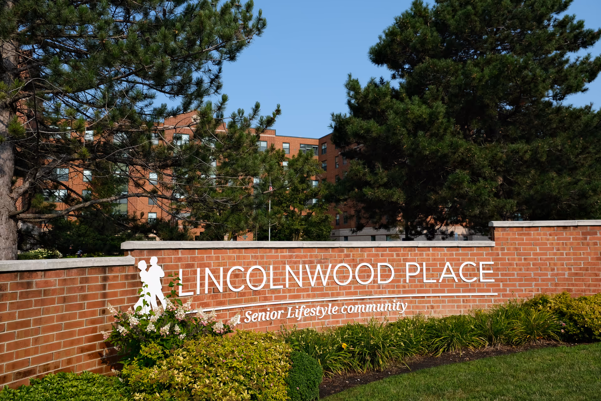 Brick wall sign for Lincolnwood Place Senior Lifestyle Community surrounded by green bushes and trees with a multi-story brick building in the background under a clear blue sky.