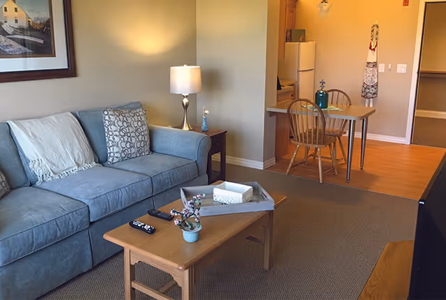 A cozy living room area with a blue sofa adorned with two pillows and a throw blanket. A wooden coffee table in front of the sofa holds a small potted plant, a tissue box, and two remote controls. To the right, there is a small dining area with a wooden table and two chairs, adjacent to a kitchen space with a refrigerator and hanging apron. A table lamp on a side table next to the sofa provides warm lighting, and a framed picture hangs on the wall above the sofa.