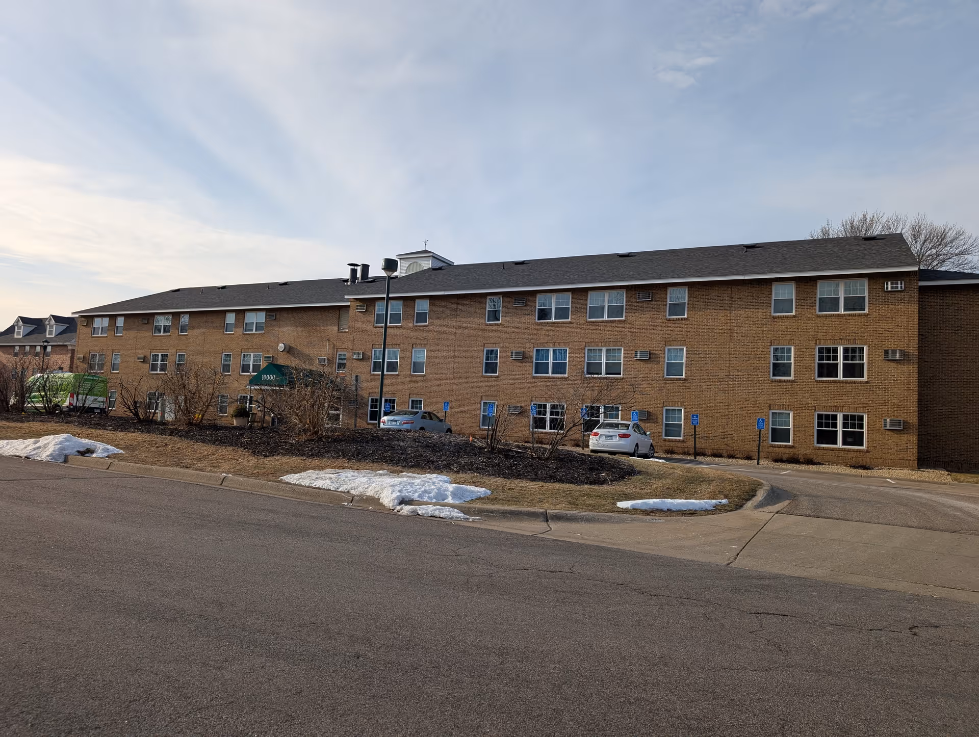 Three-story brick senior living building with a green entry canopy, parked cars, and small patches of snow on the lawn.
