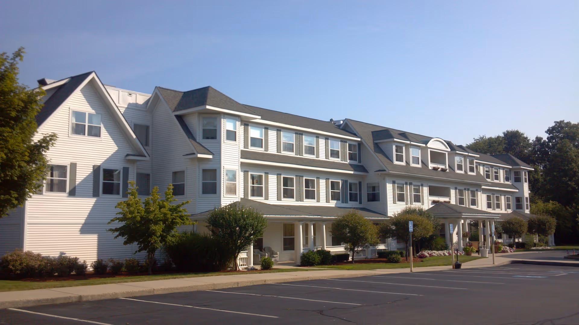 Exterior view of a large, white, multi-story senior living facility building with multiple windows and a covered entrance. The building is surrounded by small trees and bushes, with a paved parking lot in front under a clear blue sky.