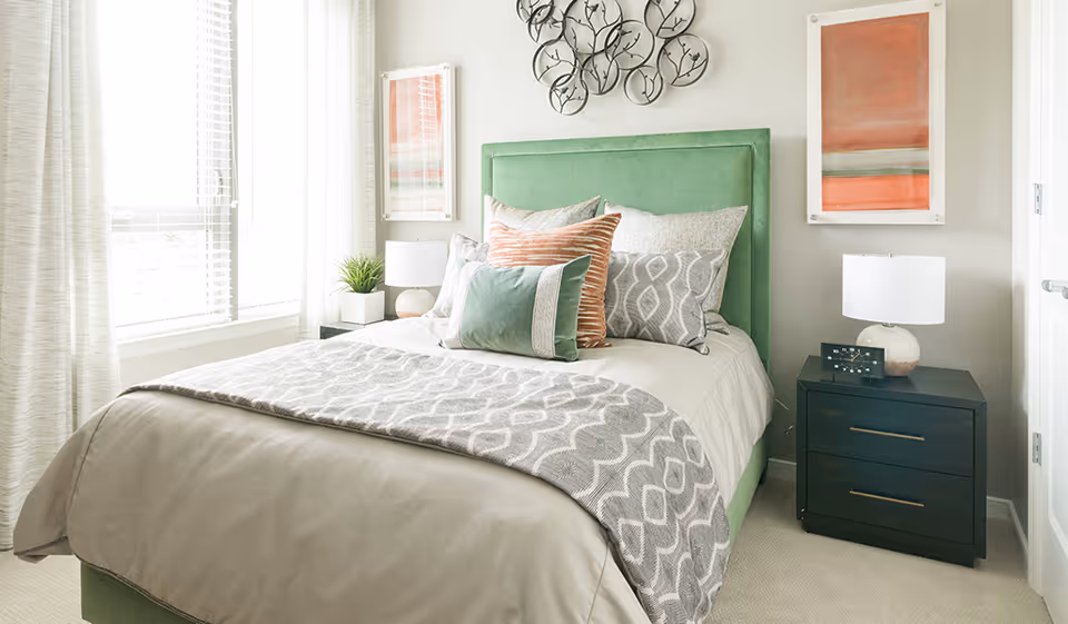 A bright bedroom featuring a bed with a green upholstered headboard, multiple decorative pillows in shades of green, gray, and orange, and a gray patterned throw blanket. There are two black nightstands on either side of the bed, each with a white lamp. The room has light-colored walls, a large window with white blinds and curtains, and two pieces of wall art above the nightstands.