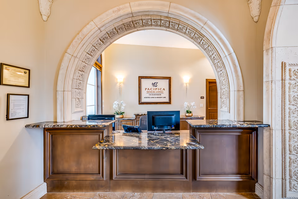 Reception desk area with a decorative stone arch overhead, dark wood counters with marble tops, two computer monitors, and a framed sign on the back wall that reads 'Pacifica Senior Living Oceanside Assisted Living & Memory Care'. Two wall sconces provide lighting and there are white orchids on the counters.