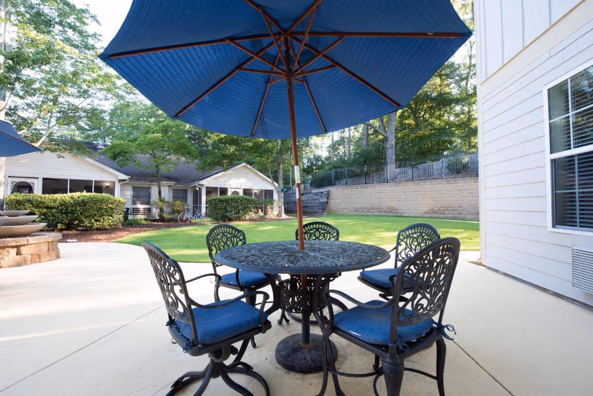 Outdoor patio area with a round metal table and four matching chairs with blue cushions under a large blue umbrella. The patio overlooks a grassy lawn with trees and a retaining wall, and there are white buildings in the background.