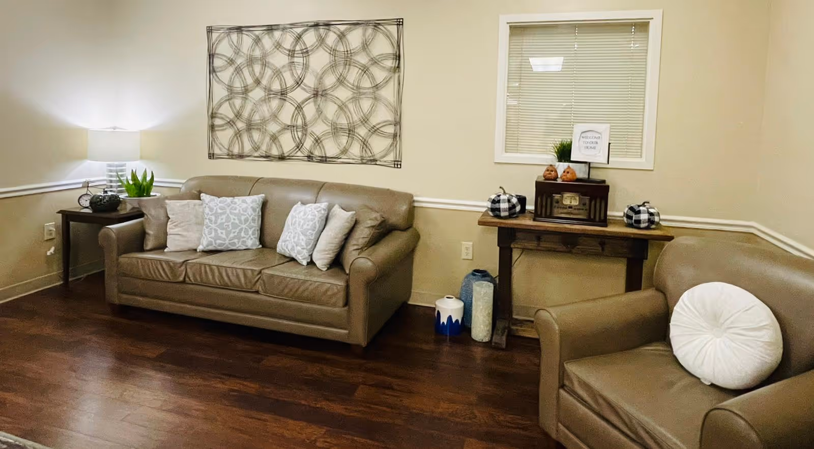 A cozy lounge with a tan leather sofa and armchair, decorative pillows, a side table with lamp, wall art, and a small console table against a beige wall.
