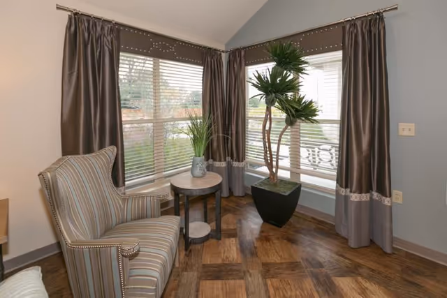 A cozy corner of a room with large windows covered by brown curtains and white blinds. There is a striped upholstered armchair next to a small round wooden table with a decorative plant on it. Another tall potted plant is placed near the windows. The floor has a wooden parquet pattern.