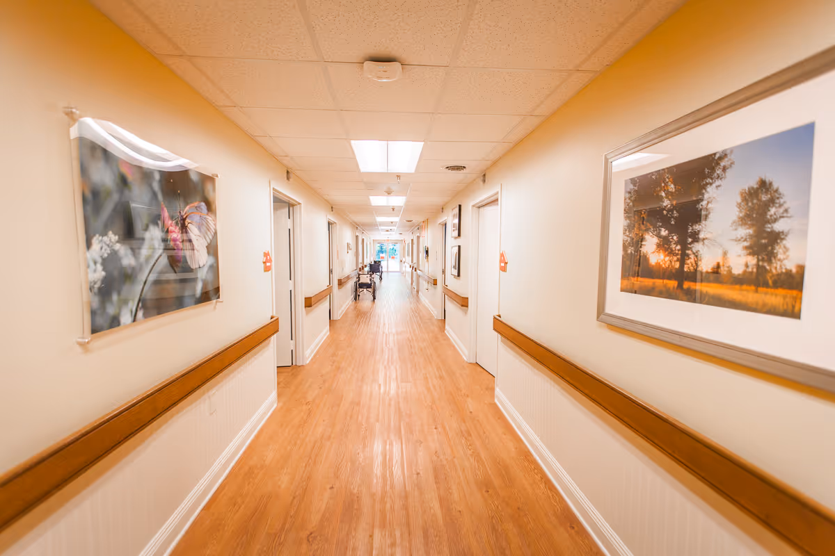 Long well-lit hallway in a nursing facility with wooden floors, handrails, and framed artwork along the walls.