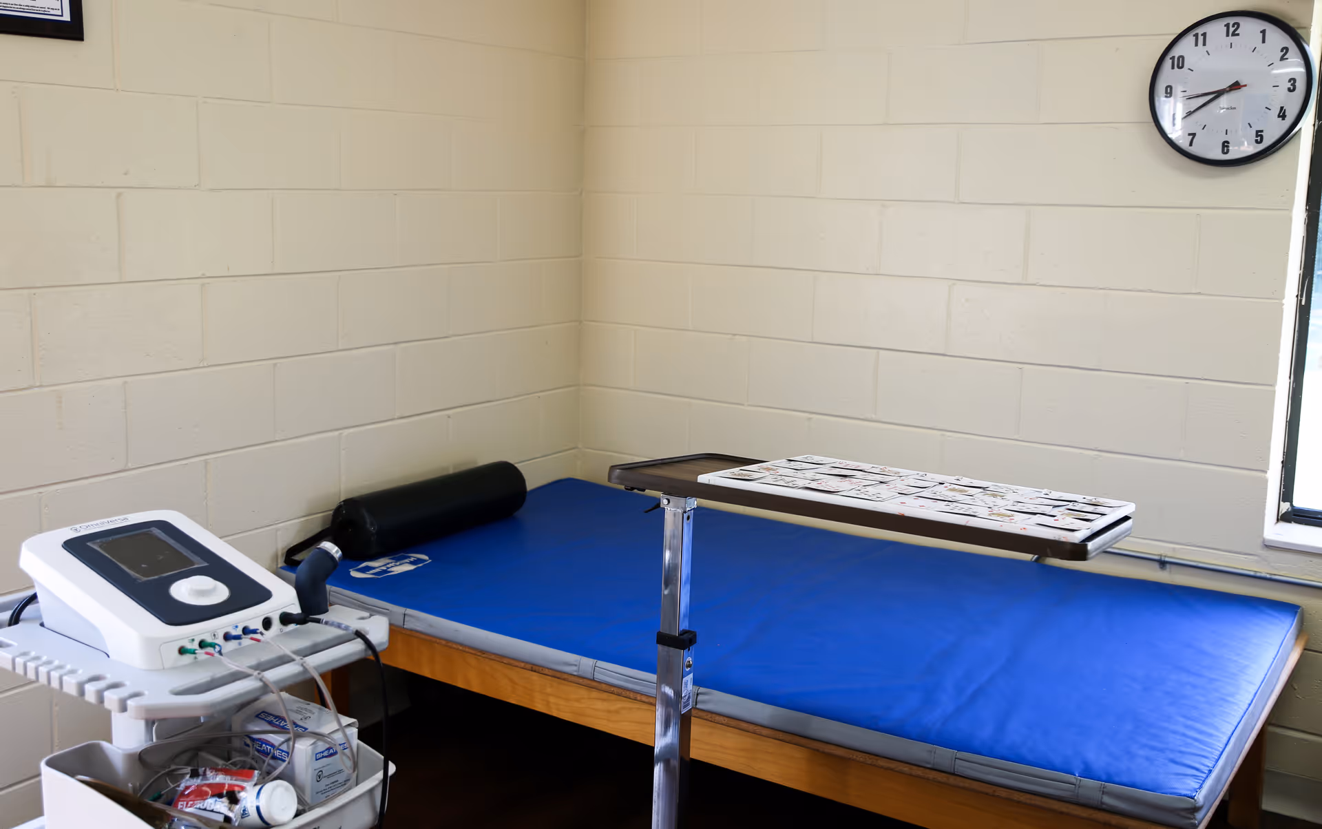 A small therapy or treatment room with a blue padded treatment table, a black cylindrical bolster pillow, a white medical device on a cart with various supplies, a wall clock showing 9:15, and a window letting in natural light.