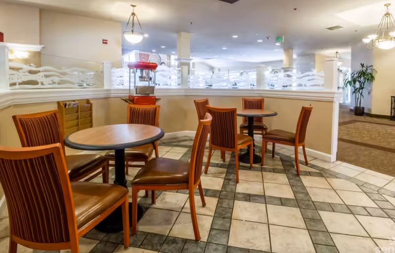 Interior view of a senior living facility dining area with round tables and wooden chairs with striped upholstery. A red popcorn machine is visible on a counter behind the tables, and decorative frosted glass panels separate the dining area from the rest of the room. The floor is tiled with a patterned design, and the space is well-lit with ceiling lights.
