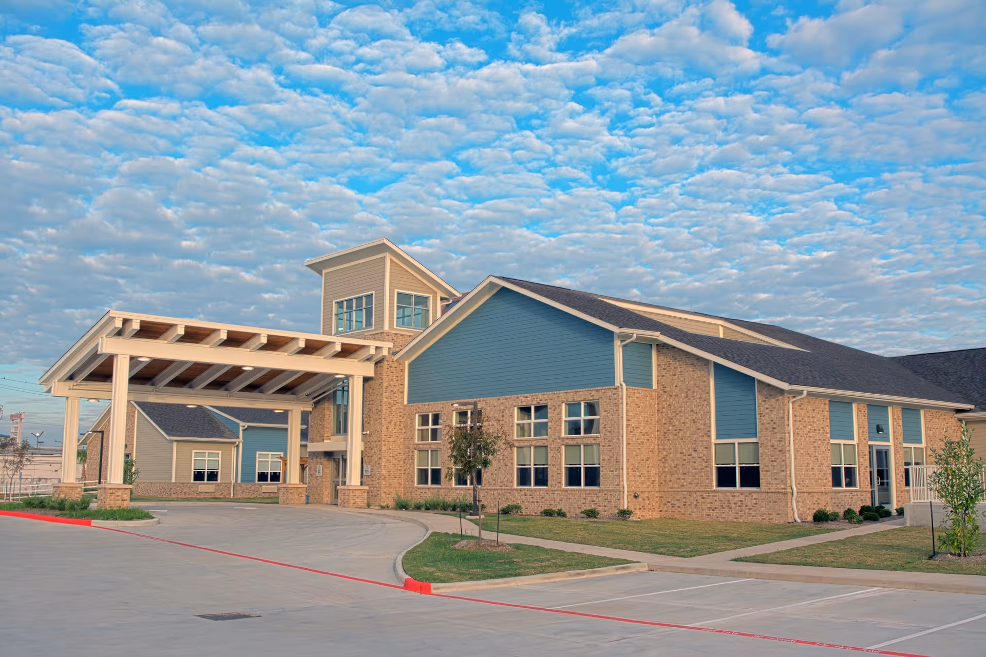 Front exterior of a brick and blue-siding senior living facility with a covered entryway under a partly cloudy sky.