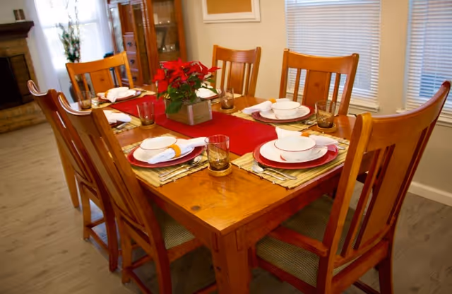 A wooden dining table set for six people with matching wooden chairs. The table is decorated with a red table runner, a centerpiece of red flowers, white bowls on red plates, folded white napkins, and drinking glasses. The room has light-colored walls, windows with blinds, and a wooden floor.
