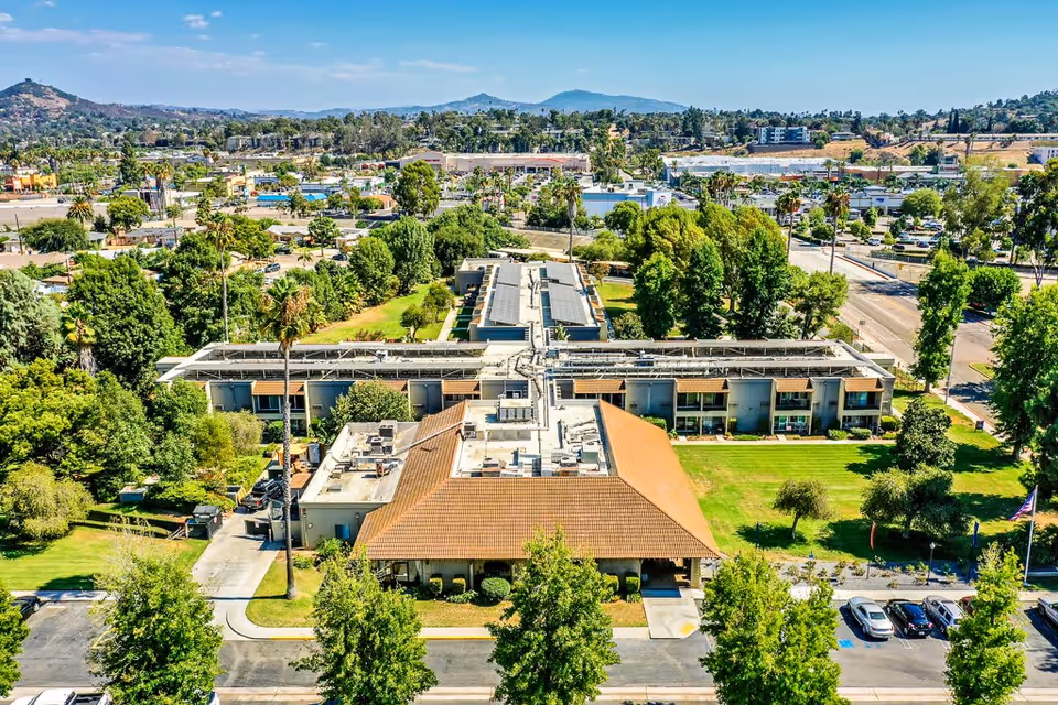 Aerial view of Escondido Senior Living facility showing a large building with a tiled roof surrounded by green lawns, trees, and parking spaces. The background includes a suburban area with more buildings, roads, and hills under a clear blue sky.