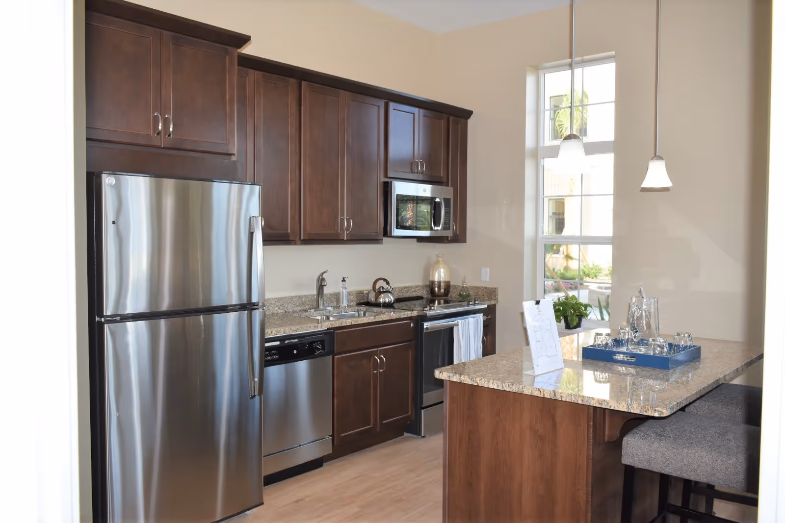 Modern kitchen with stainless steel refrigerator, dishwasher, microwave, and oven. Dark wood cabinets and granite countertops with a kitchen island featuring a tray with glasses and a water pitcher. Two pendant lights hang above the island, and a window lets in natural light.