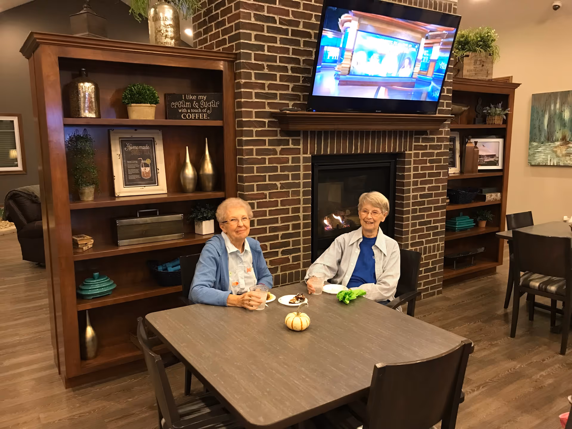Two elderly women sitting at a table in a cozy room with wooden bookshelves, a brick fireplace with a TV mounted above it, and decorative plants and items on the shelves. They are smiling and appear to be enjoying a snack and drinks.