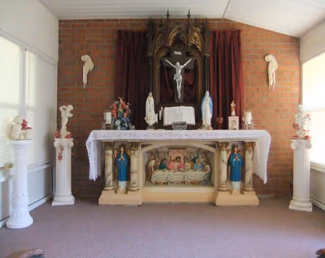 An indoor chapel altar with a crucifix, religious statues, and a lace-covered table set against a brick wall.